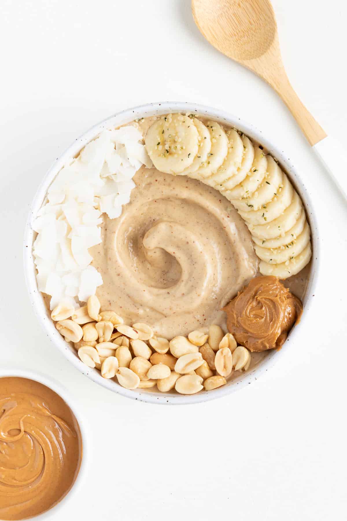 peanut butter banana smoothie bowl next to a small bowl of peanut butter and a wooden spoon