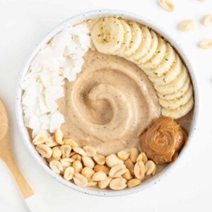 peanut butter banana smoothie bowl beside chopped peanuts and a wooden spoon