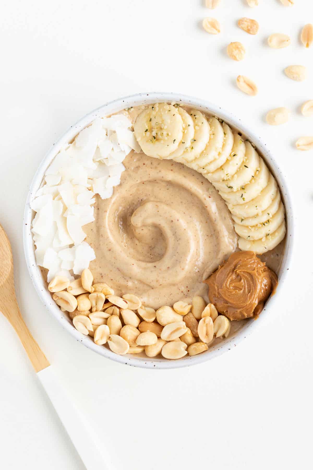 peanut butter banana smoothie bowl surrounded by chopped peanuts and a wooden spoon