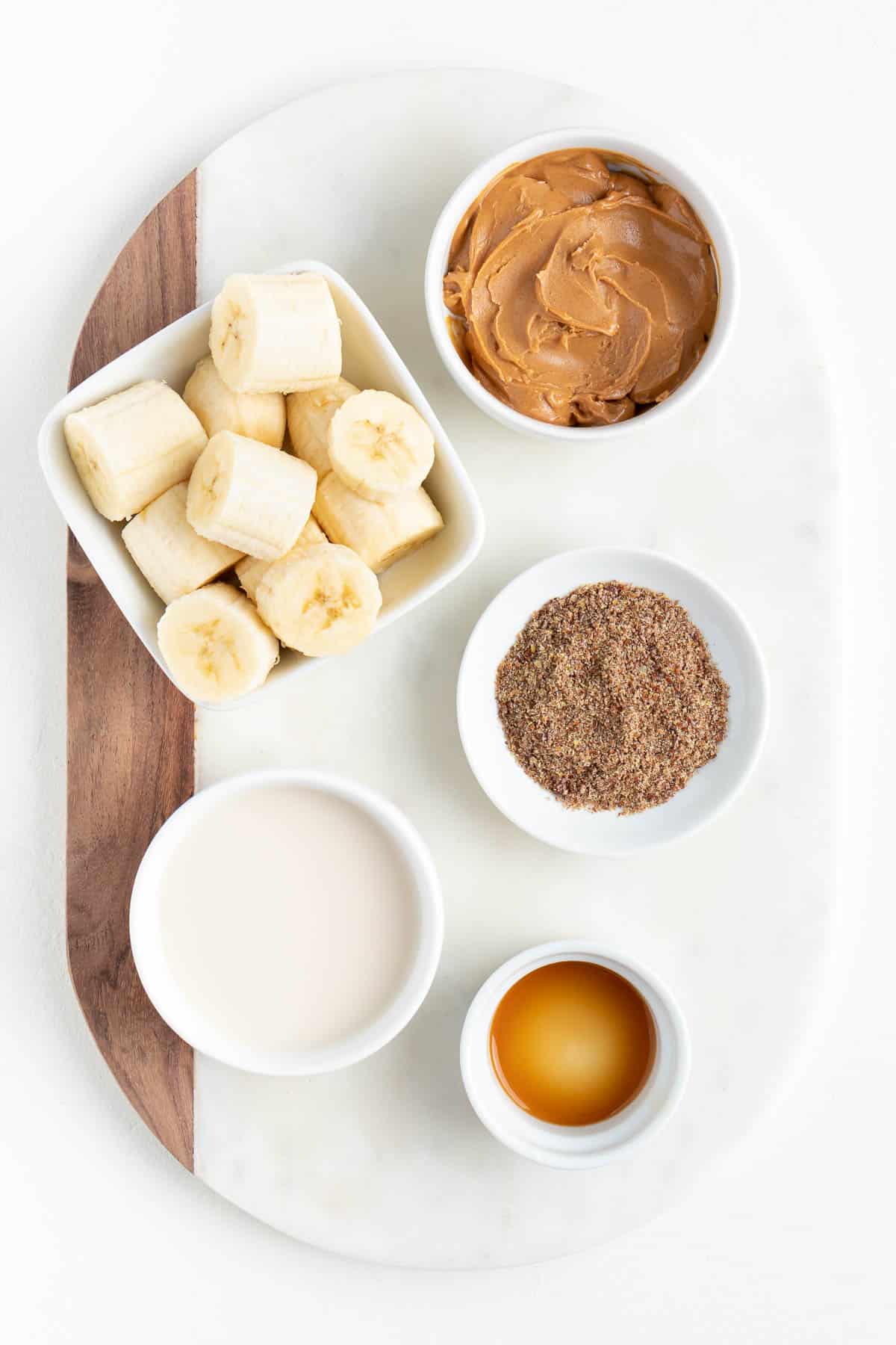 small bowls filled with chopped bananas, flaxseed, vanilla, almond milk, and nut butter on a white marble board