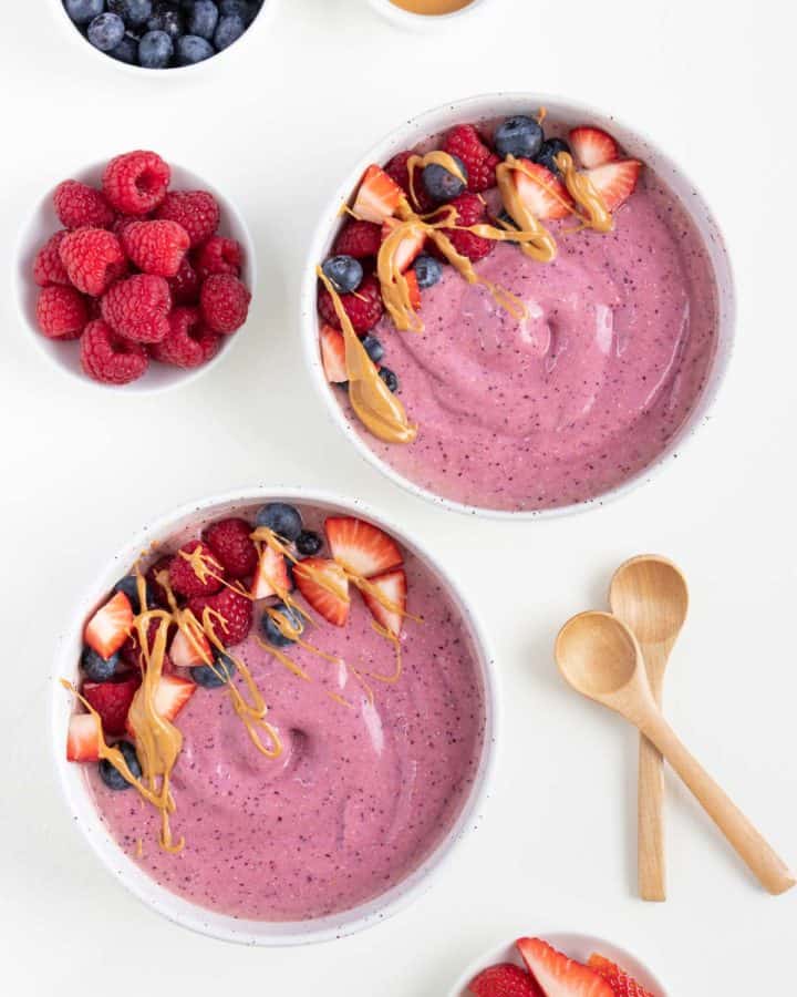 two peanut butter and jelly smoothie bowls surrounded by small white bowls filled with raspberries, blueberries, and strawberries