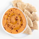 roasted red pepper hummus inside a white bowl surrounded by sliced pita bread