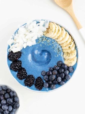a blue smoothie bowl between a bowl of blueberries and a wooden spoon
