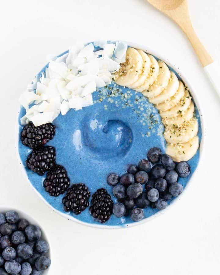 a blue smoothie bowl between a bowl of blueberries and a wooden spoon