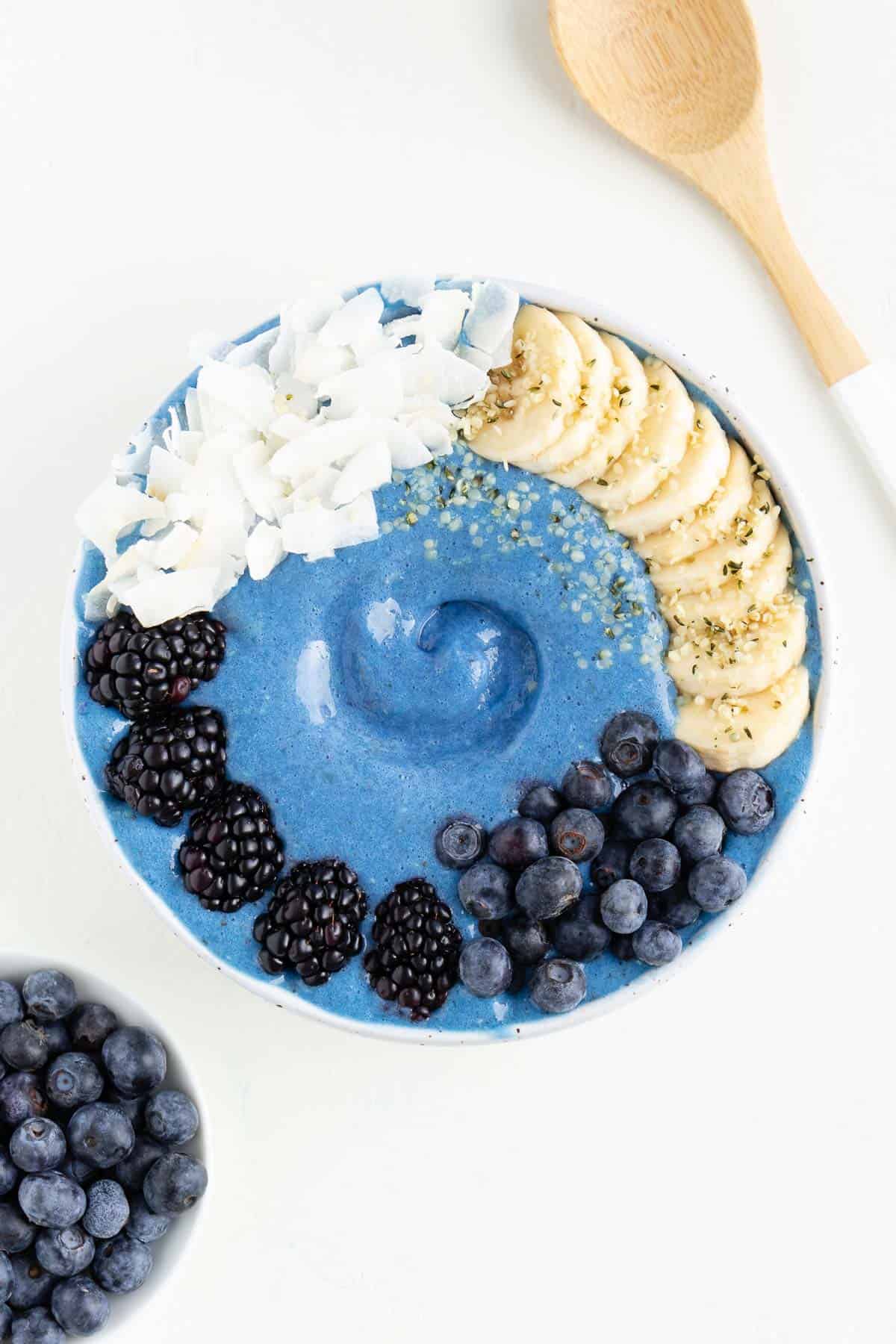 a blue smoothie bowl between a bowl of blueberries and a wooden spoon