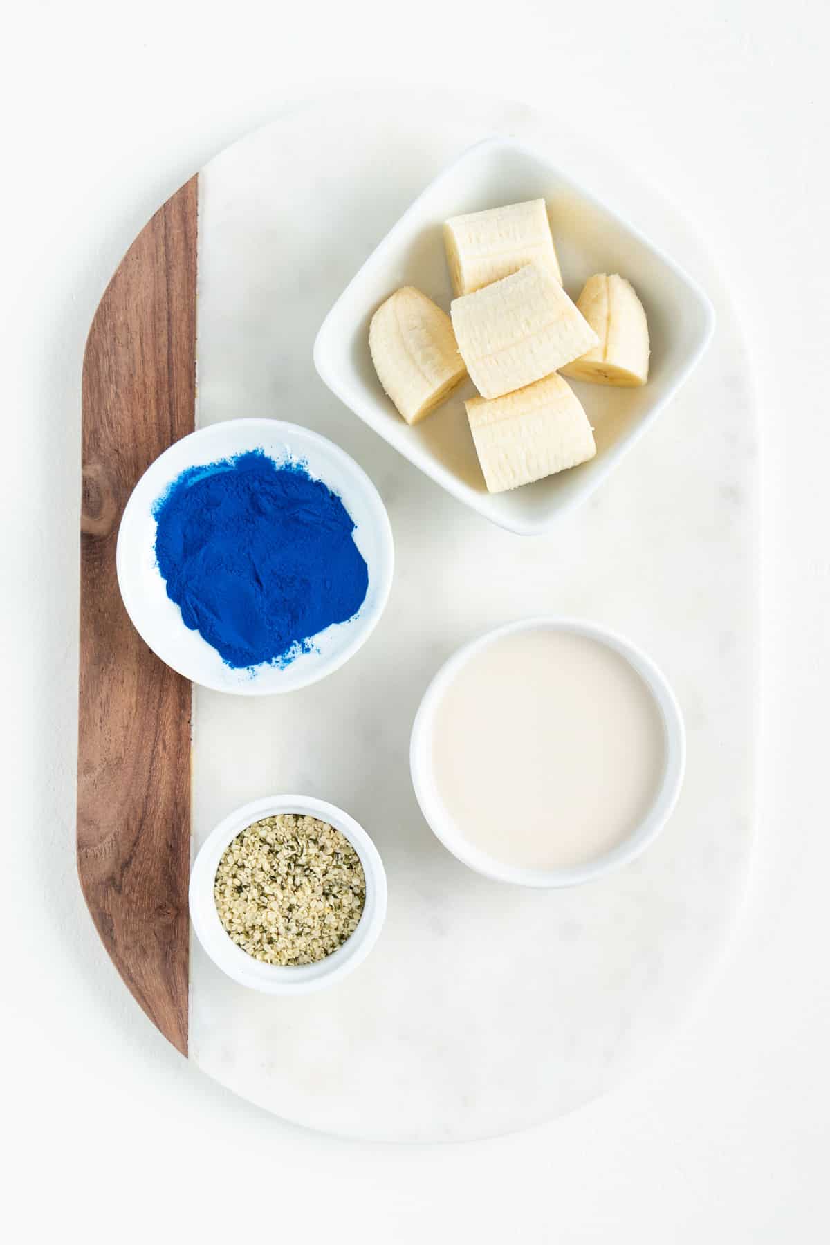 white bowls filled with bananas, spirulina powder, hemp seeds, and almond milk on a white marble board