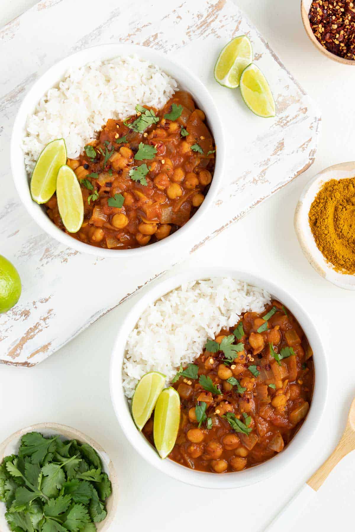 two bowls of easy chickpea curry surrounded by lime wedges and small bowls of cilantro, curry powder, and red pepper flakes