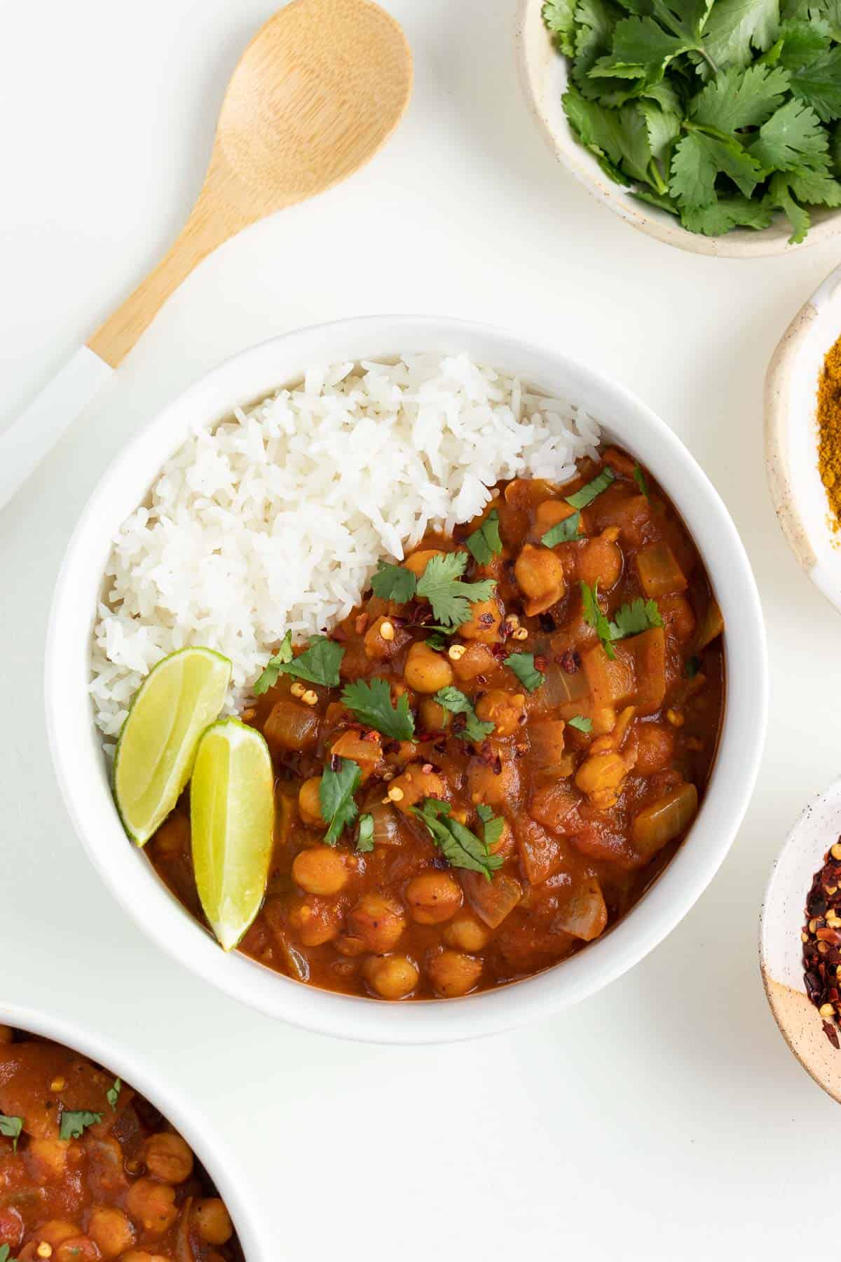 a bowl of vegan chickpea curry surrounded by cilantro, red pepper flakes, and a wooden spoon