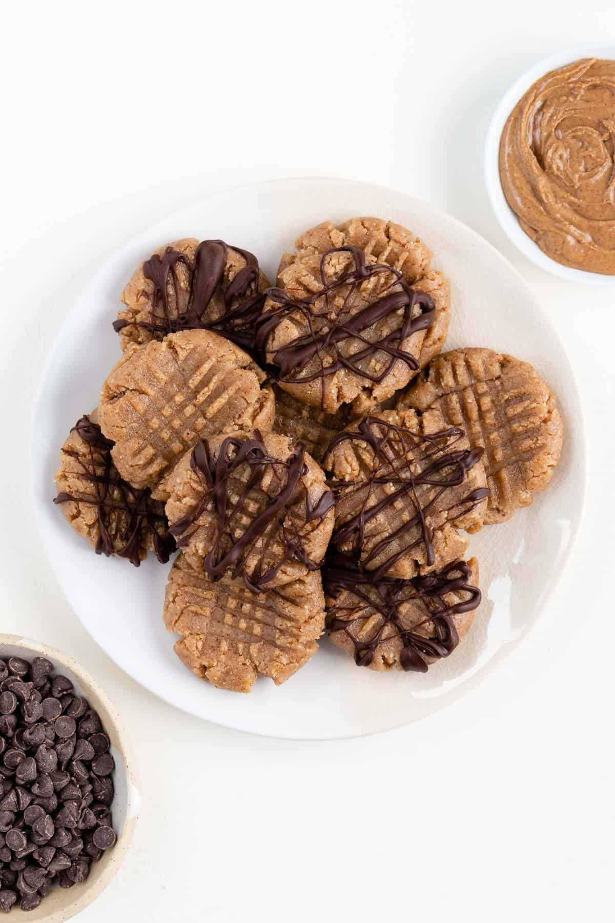 no bake almond butter cookies on a white plate surrounded by a bowl of nut butter and a bowl of chocolate chips