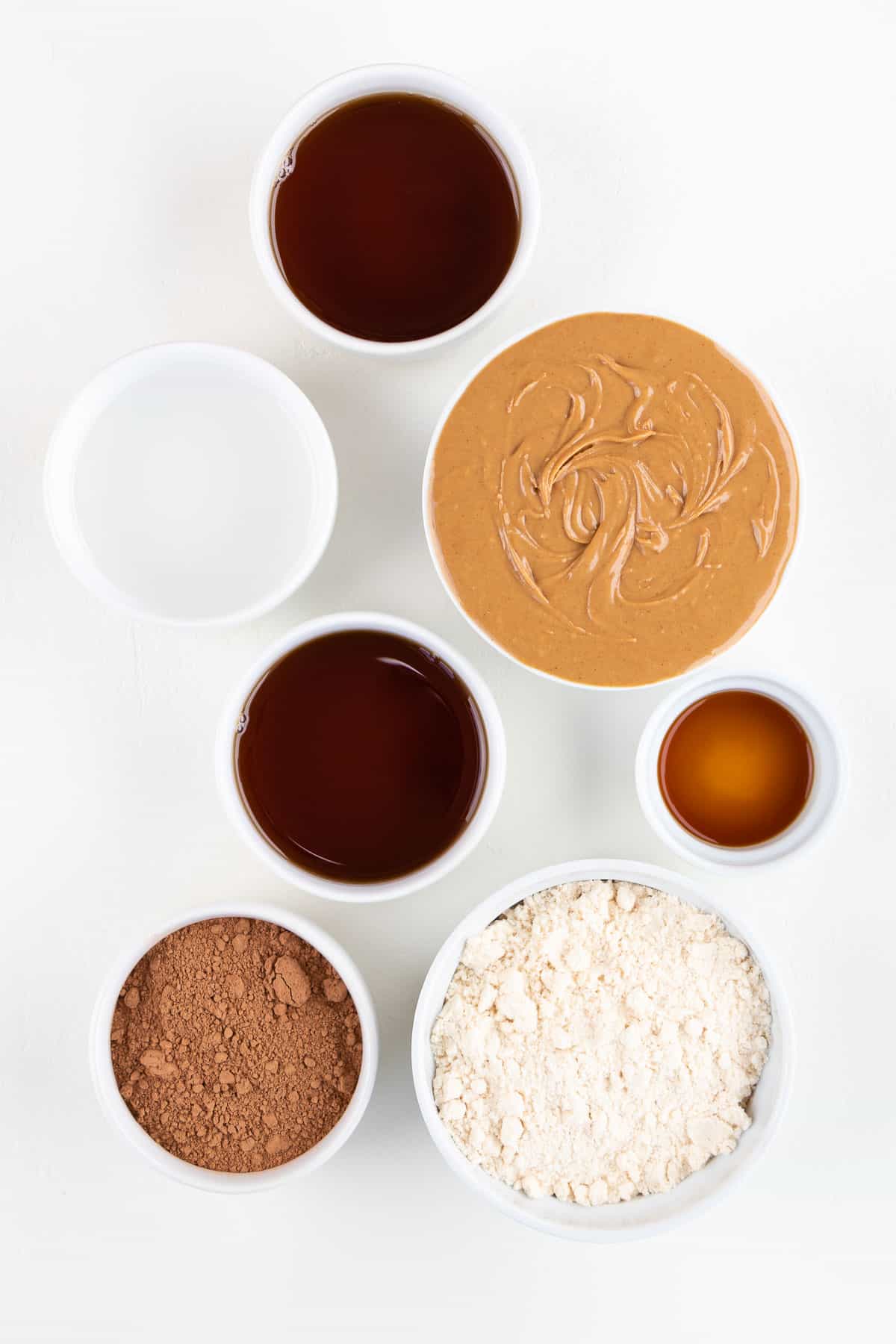 white bowls filled with coconut flour, maple syrup, nut spread, cacao powder, coconut oil, and vanilla extract