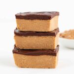 three no bake chocolate peanut butter bars stacked in front of a bowl filled with peanut butter