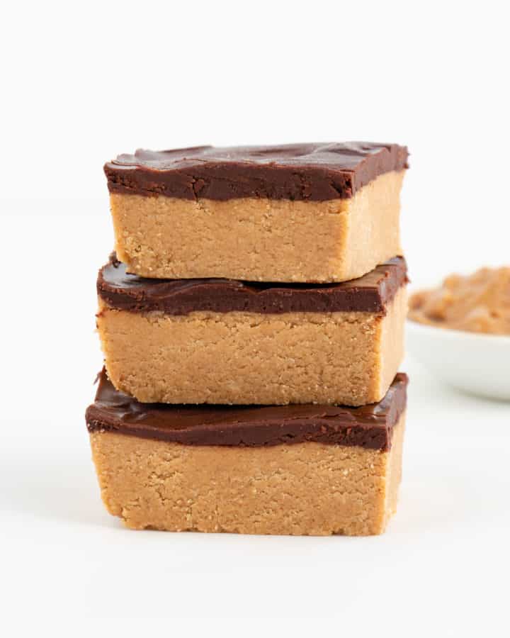 three no bake chocolate peanut butter bars stacked in front of a bowl filled with peanut butter
