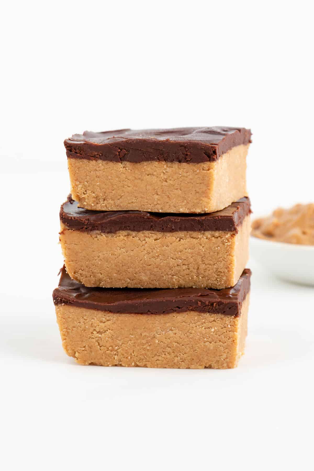 three no bake chocolate peanut butter bars stacked in front of a bowl filled with peanut butter