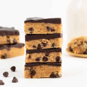 a stack of four no bake cookie dough bars stacked in front of a glass of almond milk and scoop of cookie dough on a wooden spoon