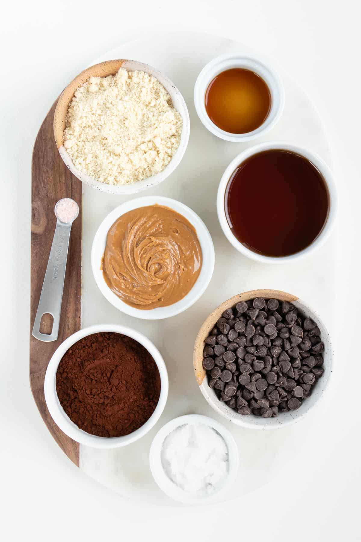 small bowls of almond flour, chocolate chips, peanut butter, cacao powder, maple syrup, vanilla, and chocolate chips on a white marble board