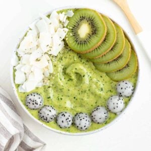 pineapple kale smoothie bowl beside a wooden spoon and grey and white striped napkin