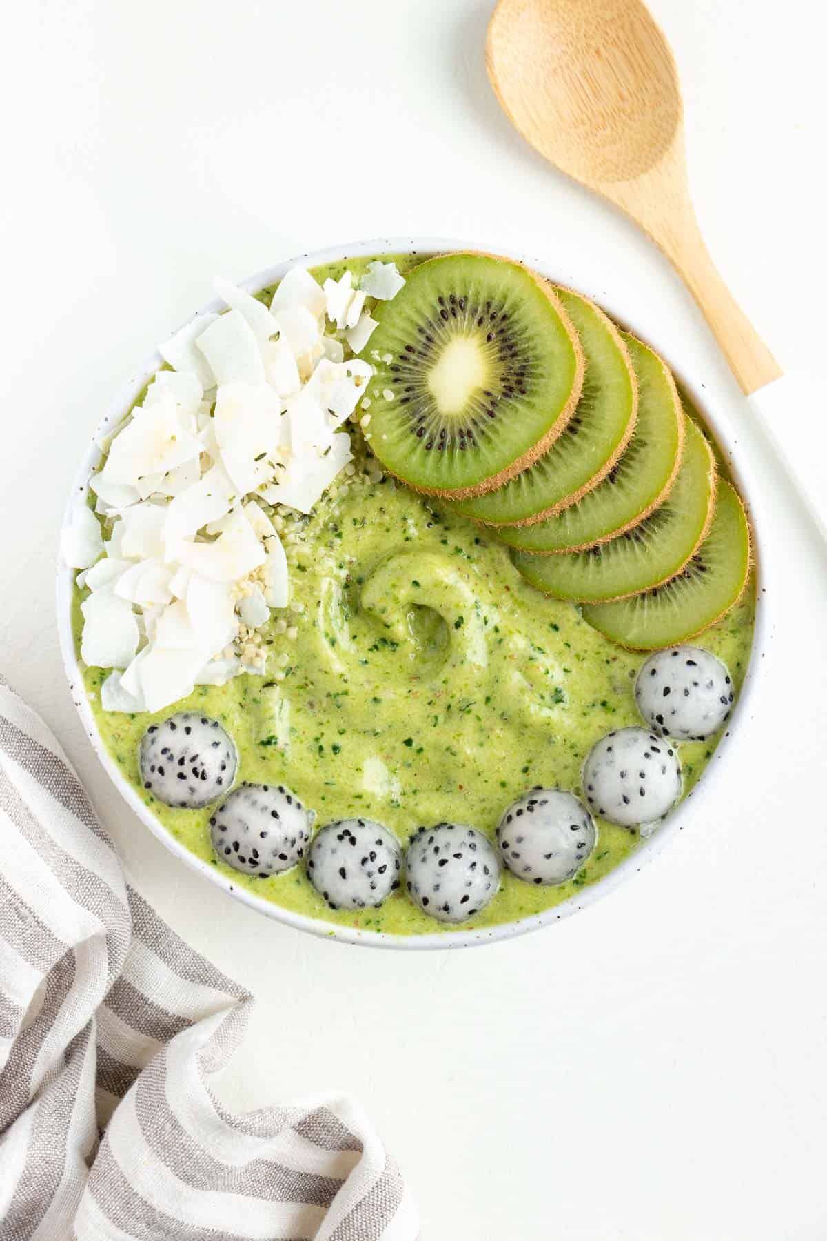 pineapple kale smoothie bowl beside a wooden spoon and grey and white striped napkin