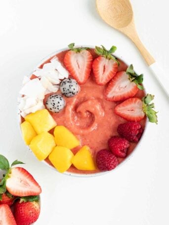 strawberry mango smoothie bowl between a wooden spoon and bowl filled with strawberries