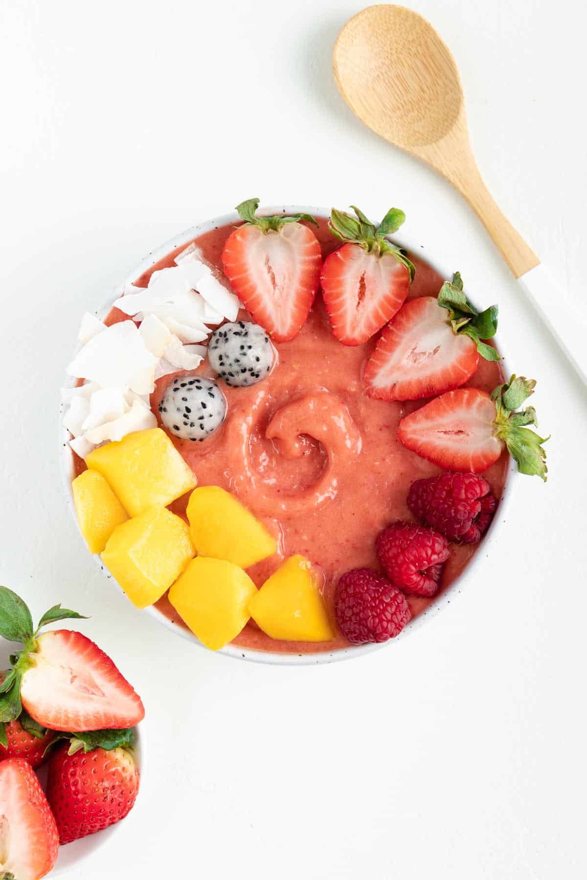 strawberry mango smoothie bowl between a wooden spoon and bowl filled with strawberries
