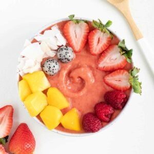 a strawberry mango smoothie bowl between a wooden spoon and sliced strawberries