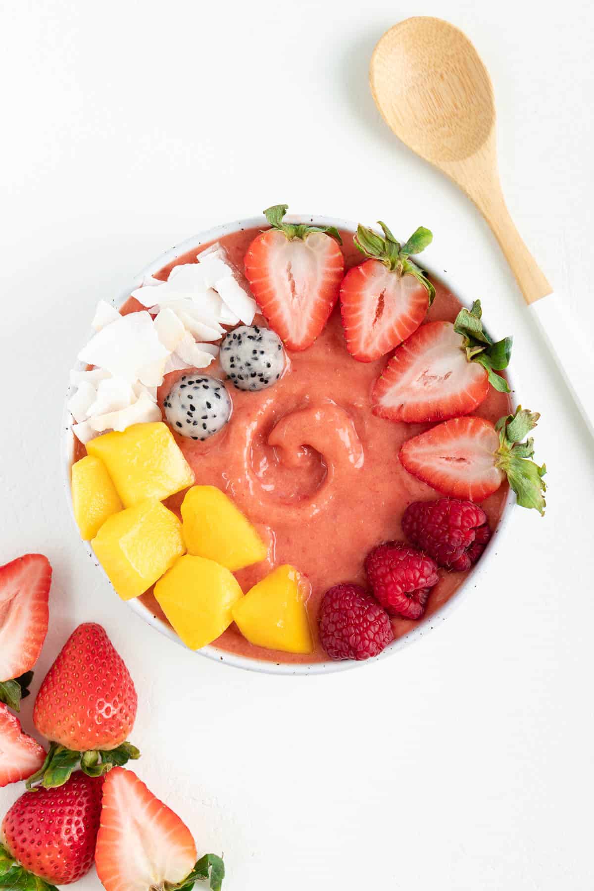 a strawberry mango smoothie bowl between a wooden spoon and sliced strawberries