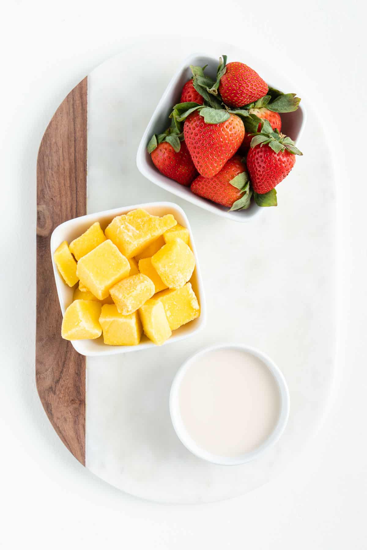 two bowls of fresh fruit and a glass of almond milk on top of a white marble board