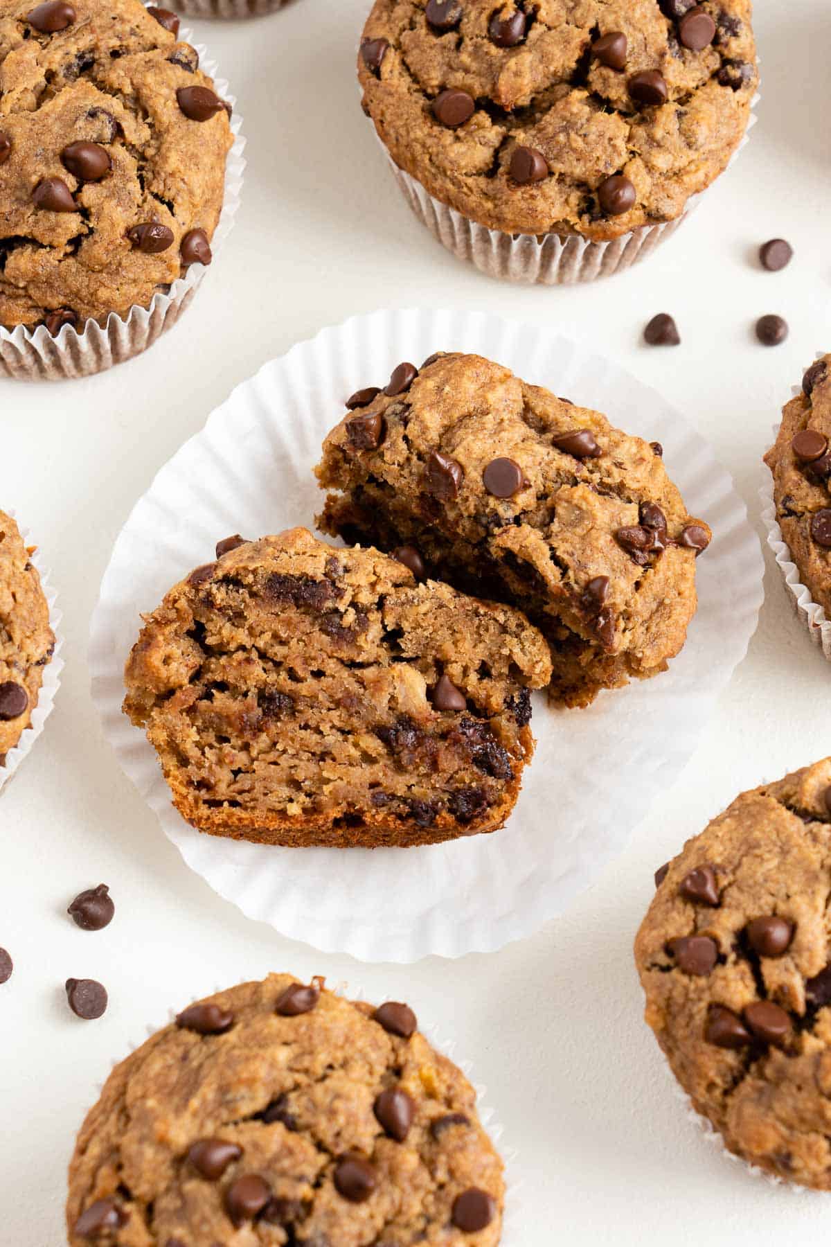 a vegan banana chocolate chip muffin sliced in half laying on a white muffin liner surrounded by more muffins