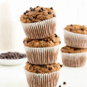 three gluten-free vegan banana chocolate chip muffins stacked on top of each other in front of a glass of almond milk and bowl of chocolate