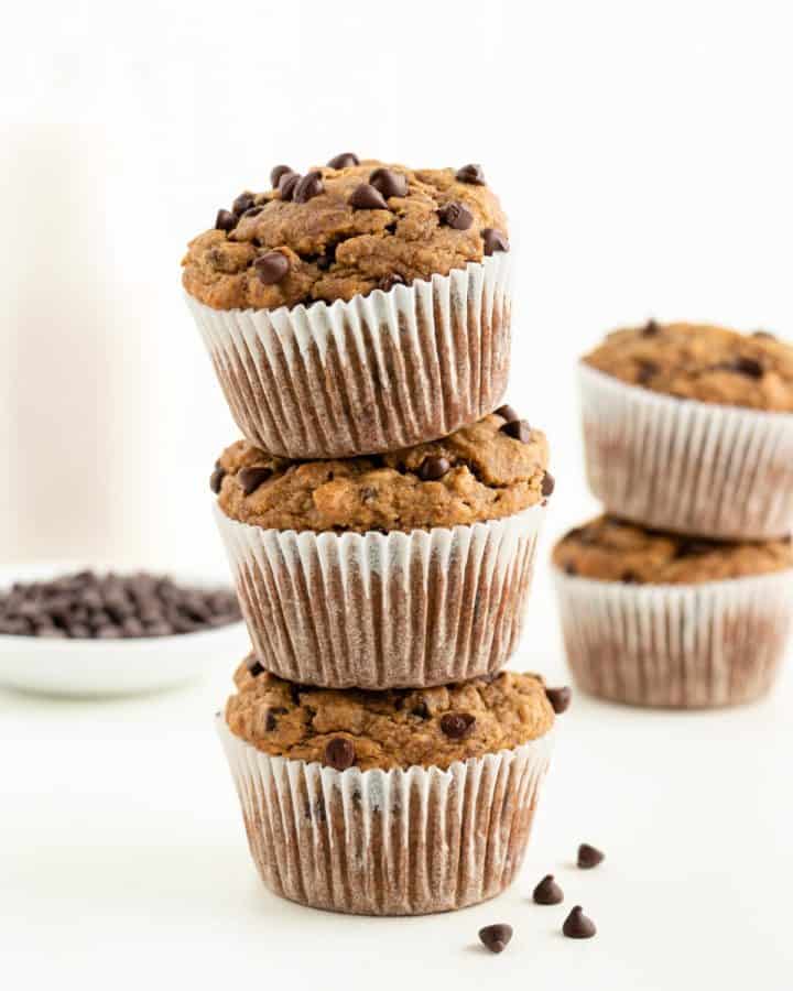 three gluten-free vegan banana chocolate chip muffins stacked on top of each other in front of a glass of almond milk and bowl of chocolate