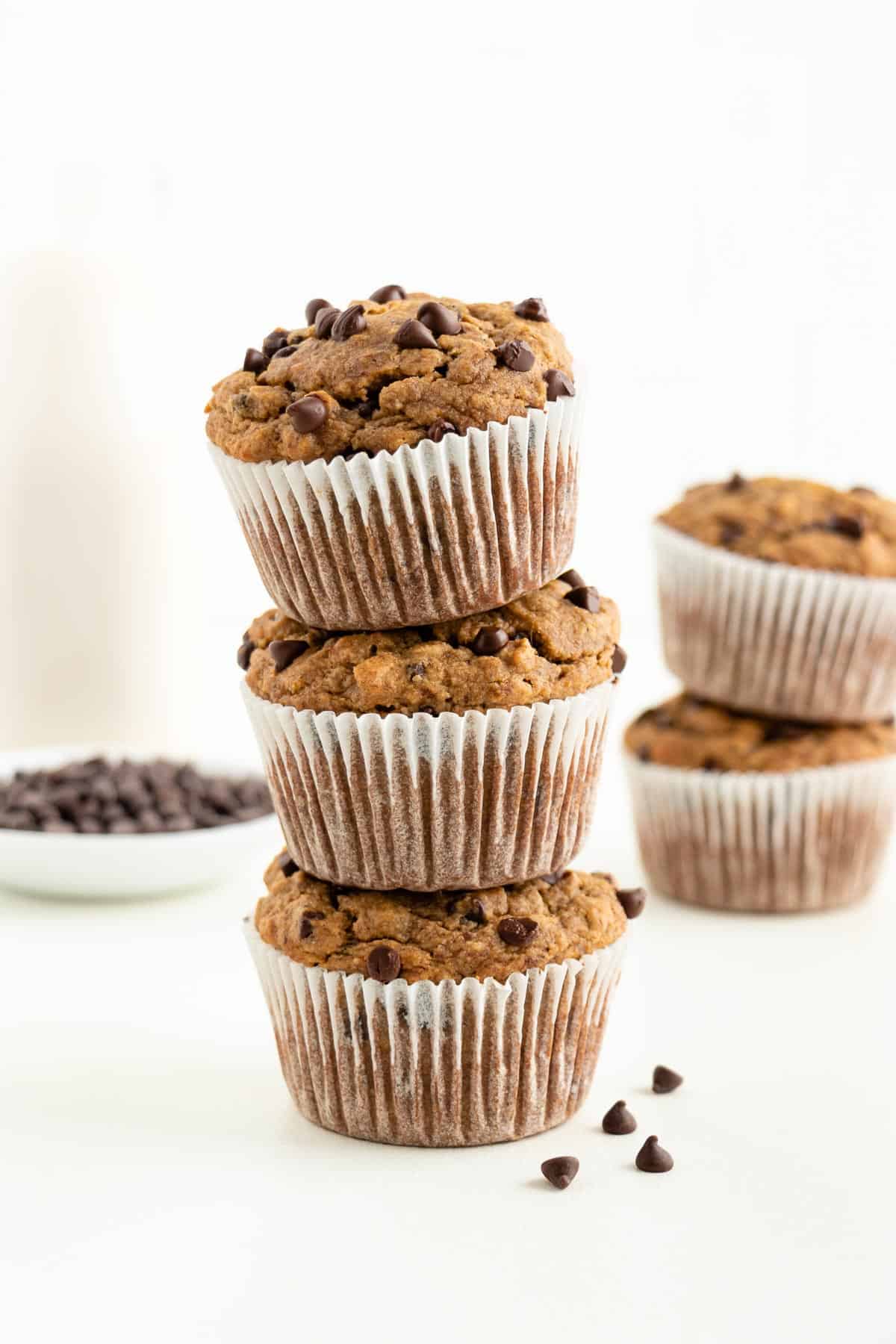 three gluten-free vegan banana chocolate chip muffins stacked on top of each other in front of a glass of almond milk and bowl of chocolate