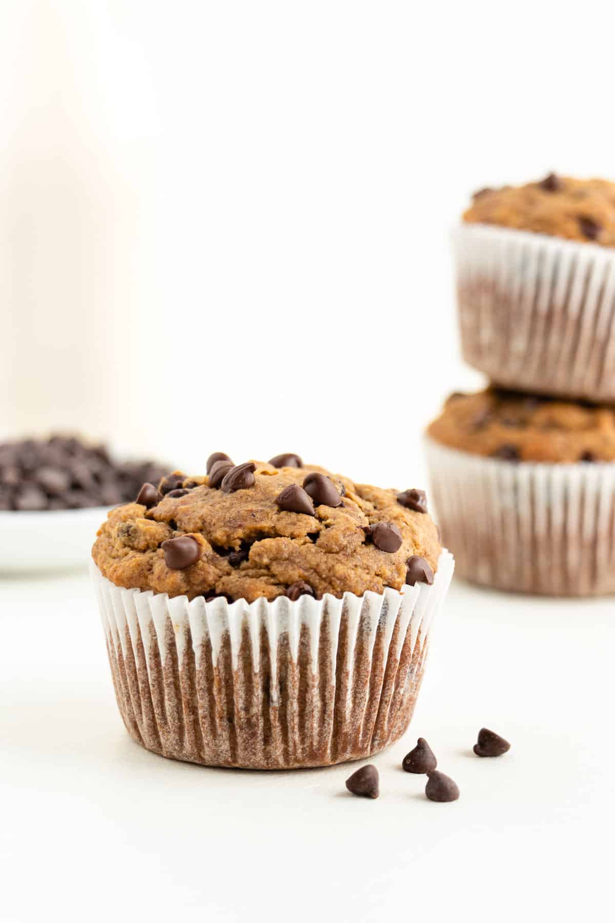 a gluten-free vegan chocolate chip muffin in front of a glass of almond milk and bowl of chocolate chips