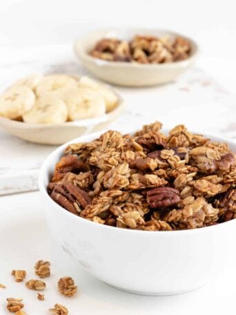 banana bread granola in a white bowl with sliced bananas and walnuts behind it