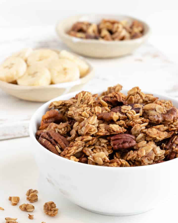 banana bread granola in a white bowl with sliced bananas and walnuts behind it