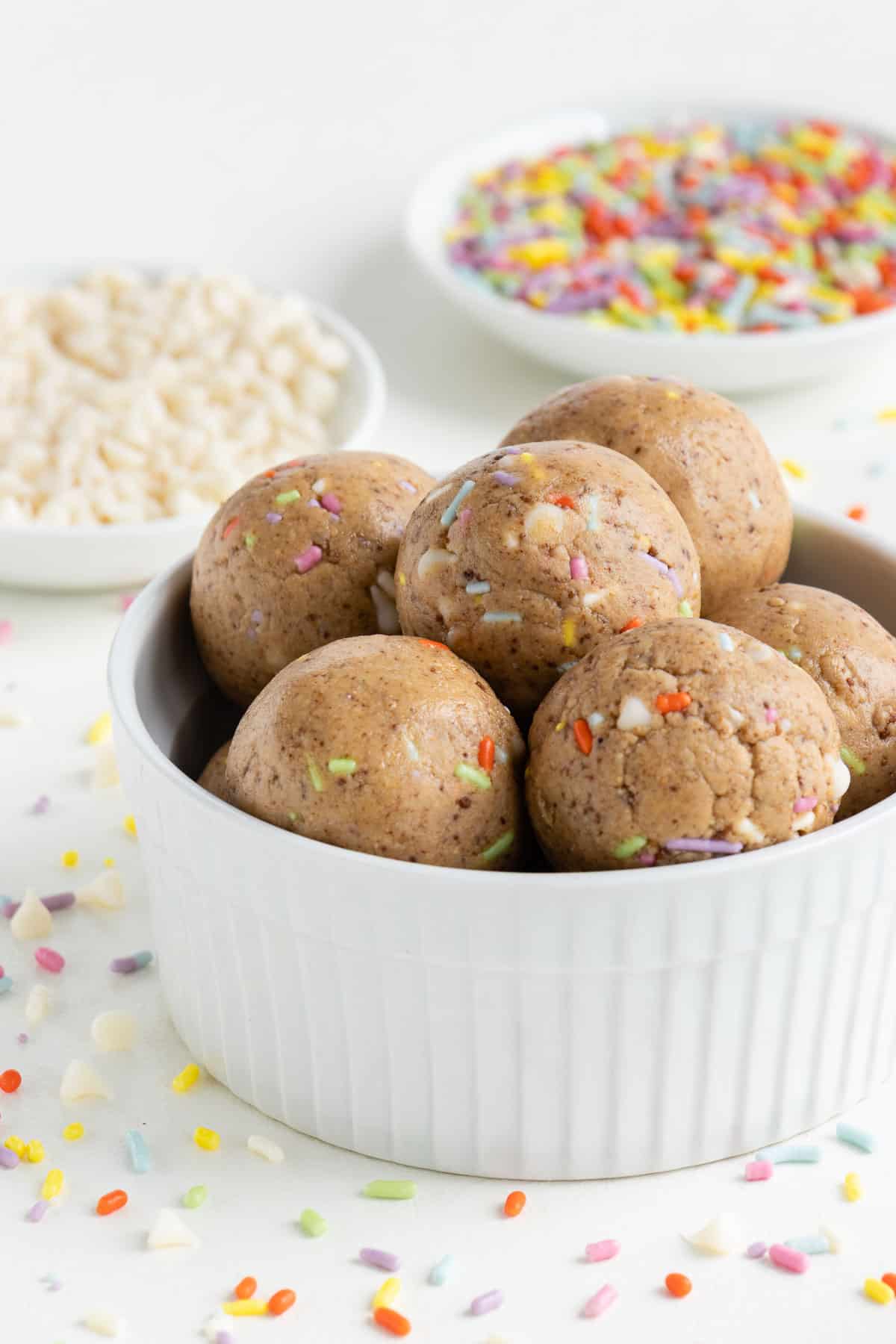 birthday cake energy balls stacked inside a white bowl beside white chocolate chips and sprinkles