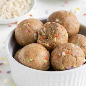 birthday cake energy balls stacked inside a white bowl beside white chocolate chips and sprinkles