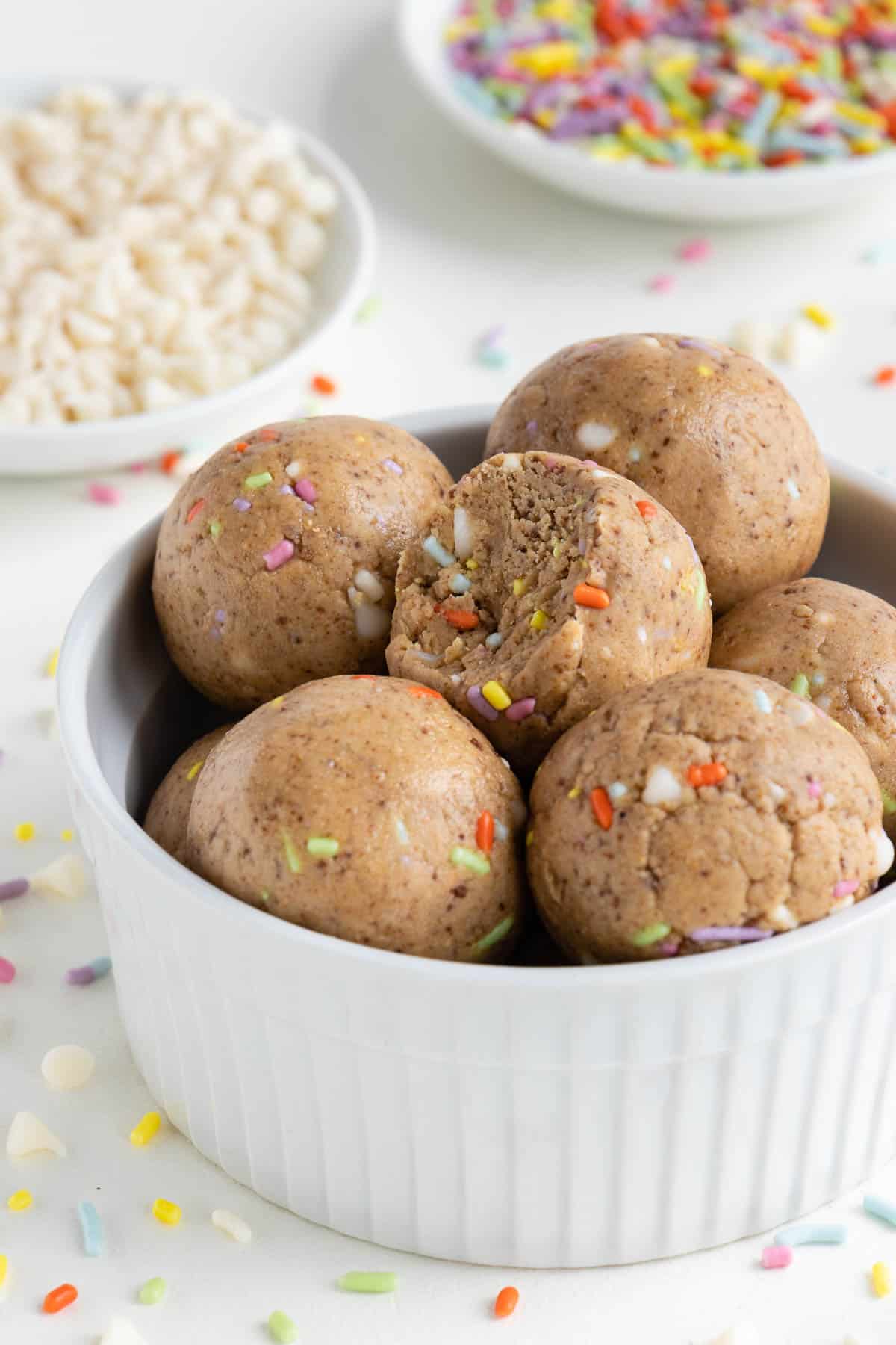 birthday cake energy balls stacked inside a white bowl beside white chocolate chips and sprinkles