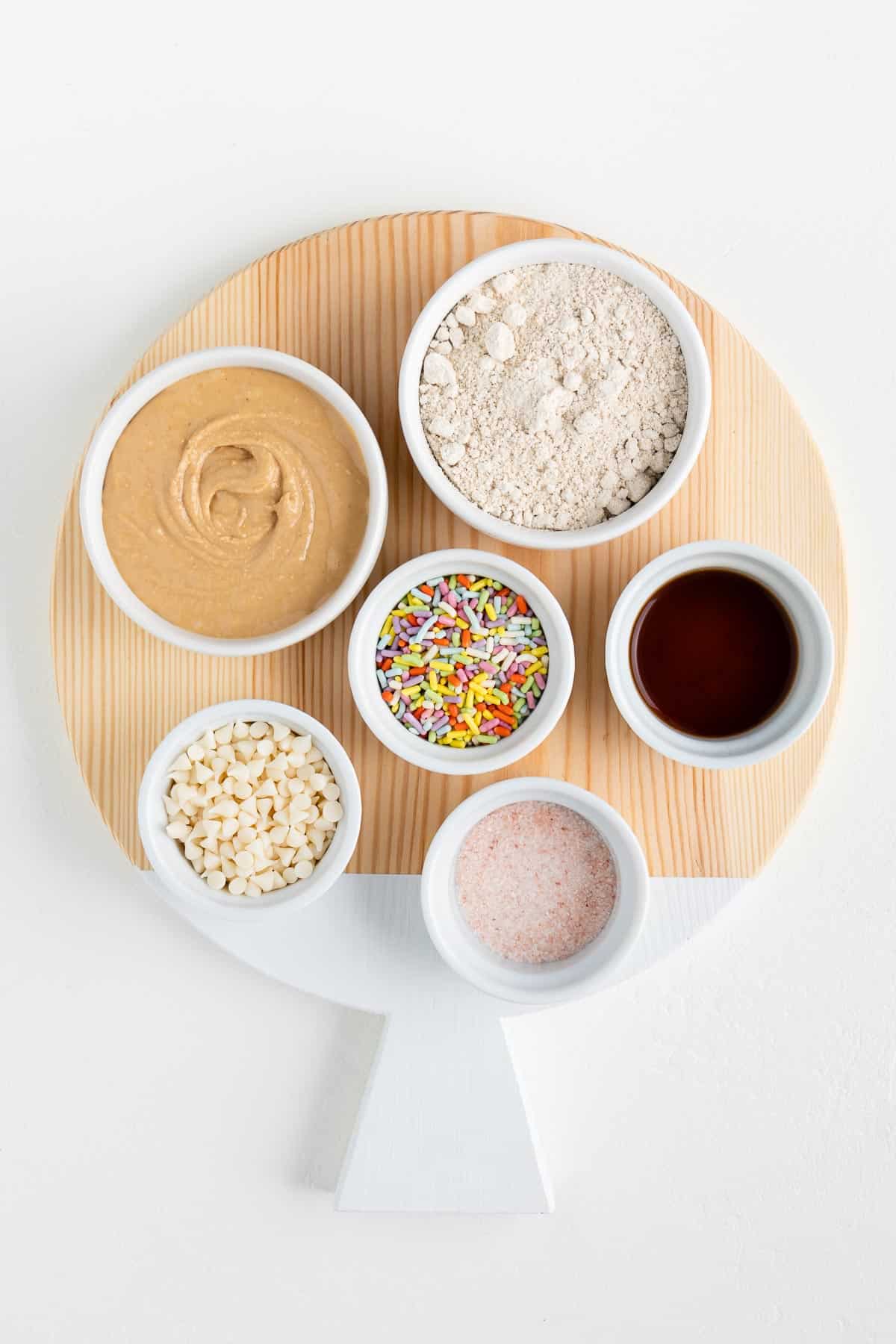 round wooden cutting board with cashew butter, sprinkles, oat flour, vanilla extract, and white chocolate chips in small white bowls