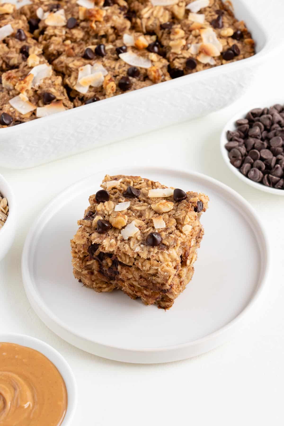 a square of chunky monkey baked oatmeal on a white plate surrounded by a small bowl of chocolate chips