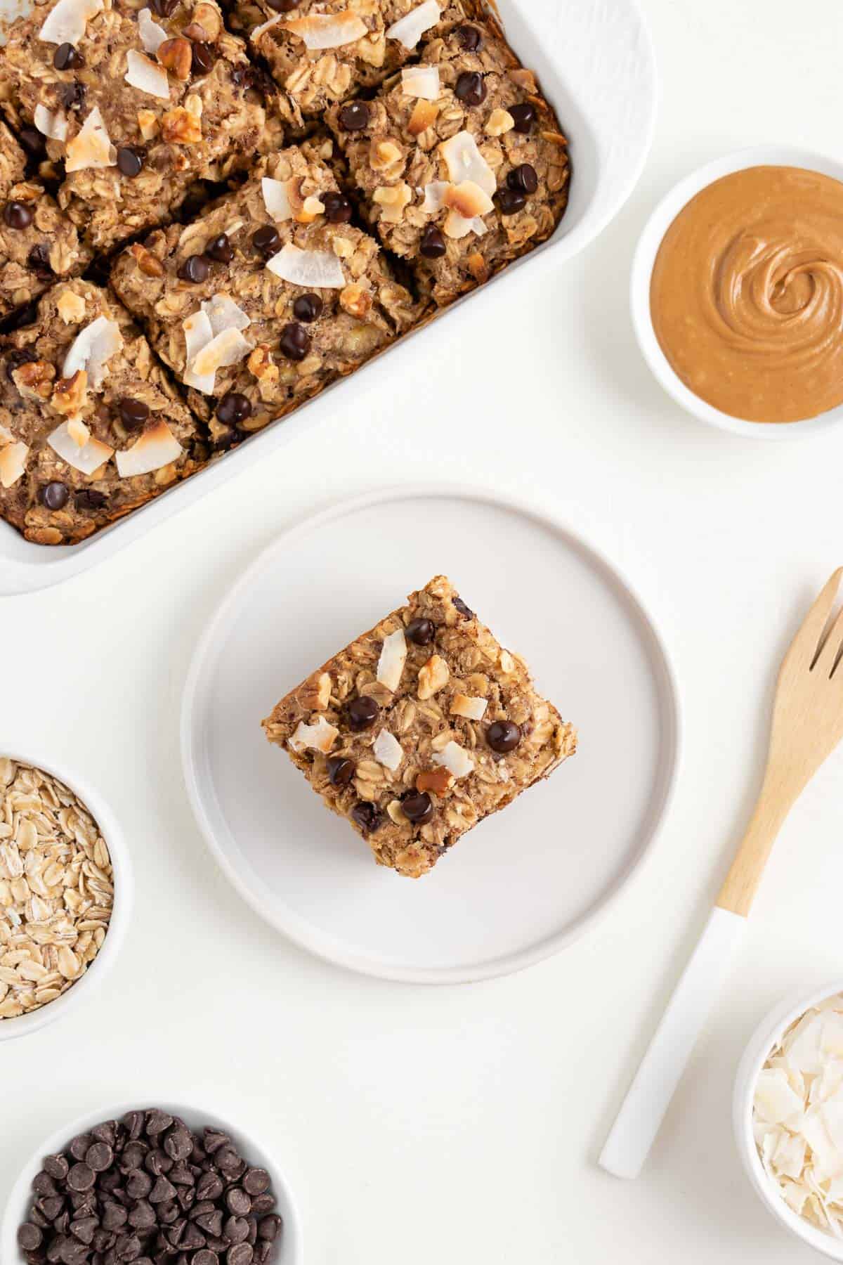 chocolate chip banana baked oatmeal on a white plate beside bowls of peanut butter, oats, chocolate chips, and coconut