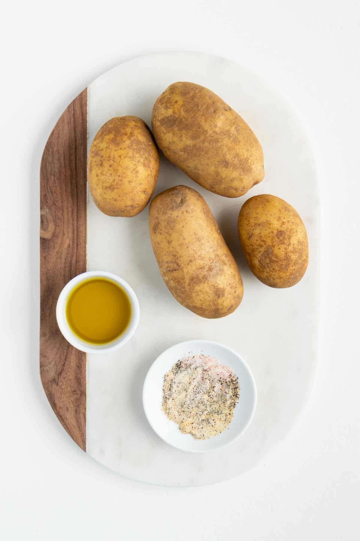 four russett potatoes, a bowl of olive oil, and a bowl of seasoning on a white marble board