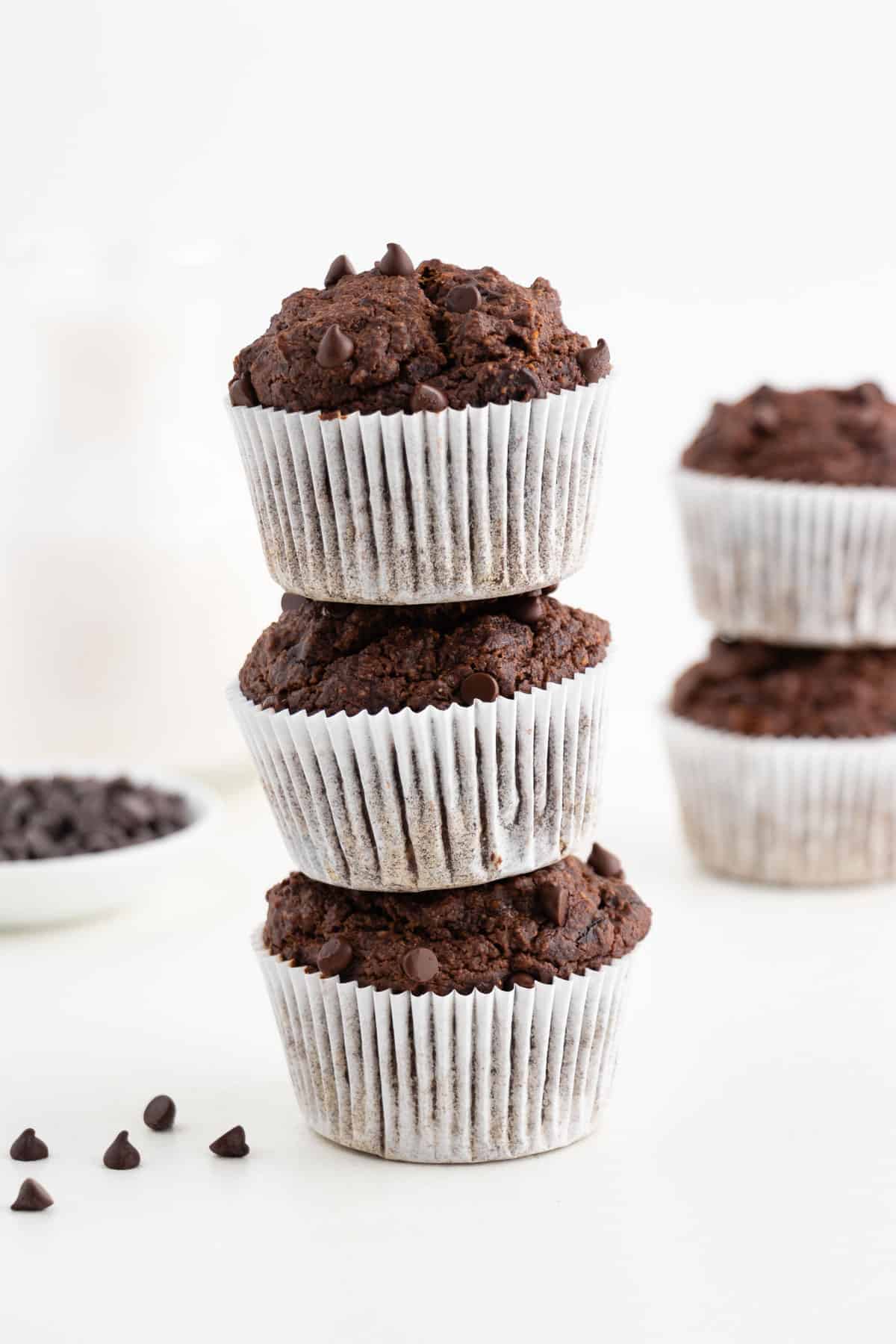 a stack of three vegan gluten-free double chocolate banana muffins in front of a glass of almond milk and bowl of chocolate chips