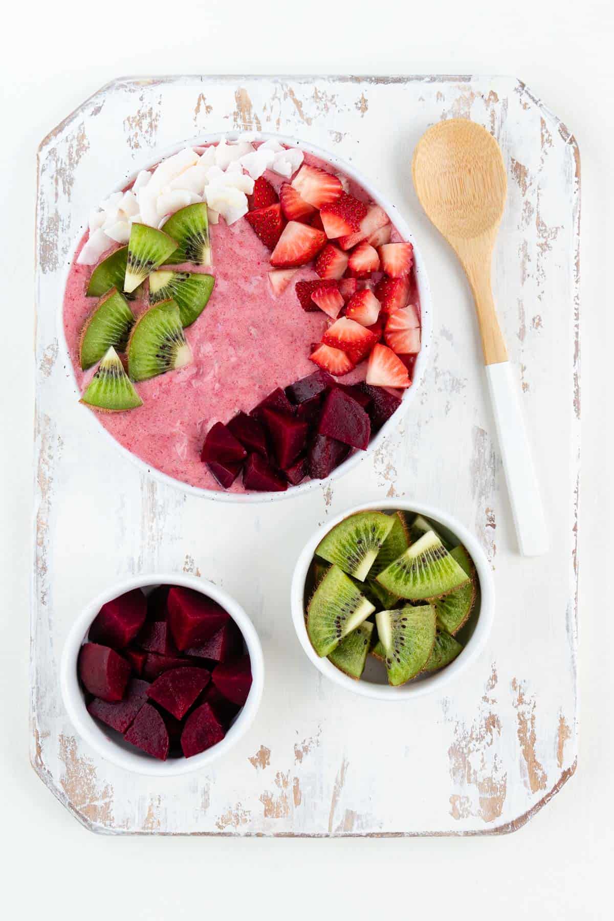 a pink smoothie bowl, wooden spoon, and two bowls filled with beets and kiwi on top of a white distressed cutting board