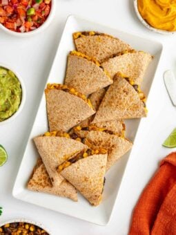 sweet potato black bean quesadillas on a white plate surrounded by bowls of guacamole, salsa, pico de gallo, and cilantro