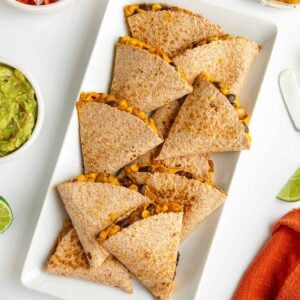 sweet potato black bean quesadillas on a white plate surrounded by bowls of guacamole, salsa, pico de gallo, and cilantro