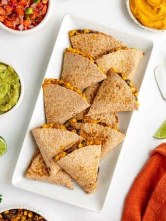 sweet potato black bean quesadillas on a white plate surrounded by bowls of guacamole, salsa, pico de gallo, and cilantro