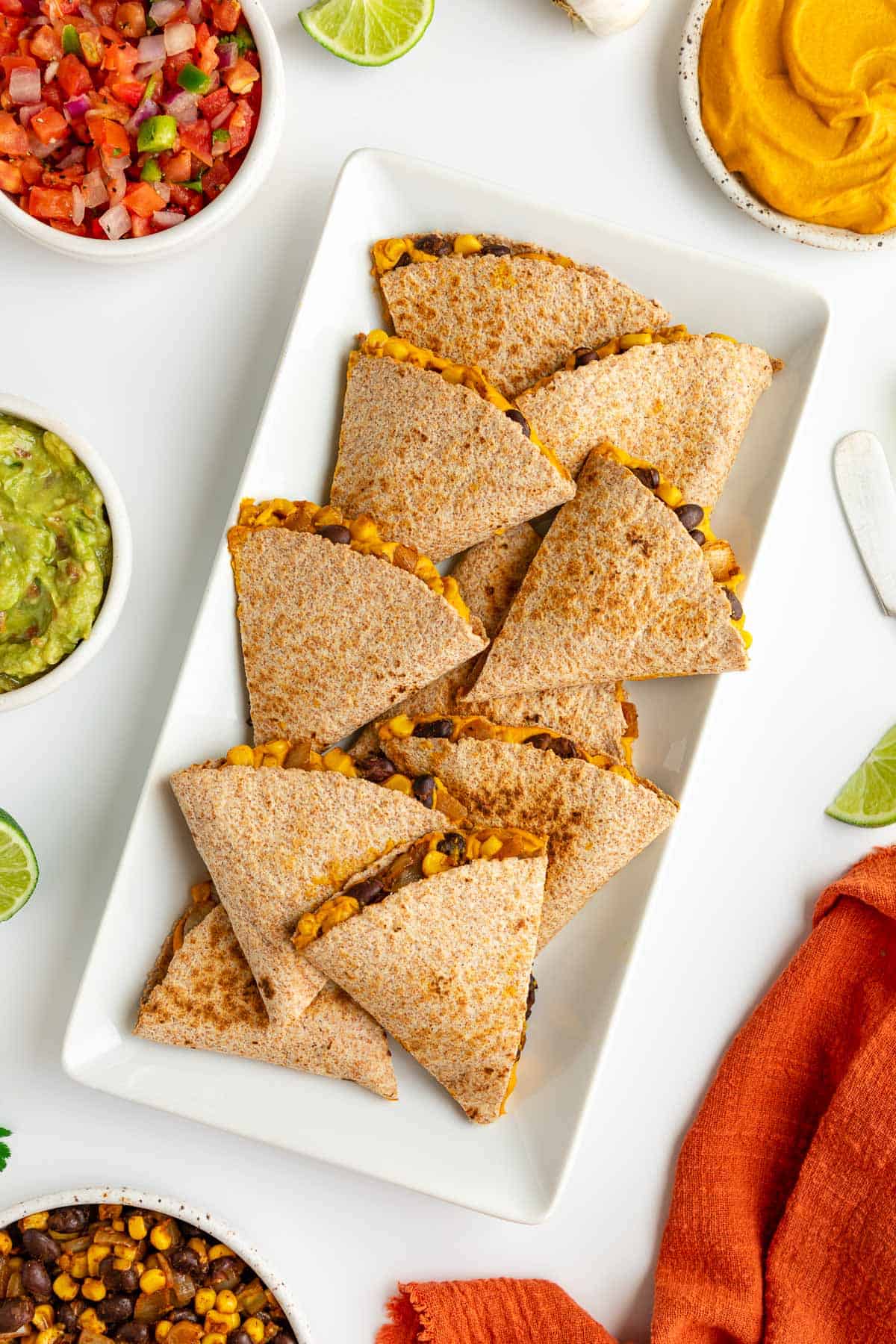 sweet potato black bean quesadillas on a white plate surrounded by bowls of guacamole, salsa, pico de gallo, and cilantro