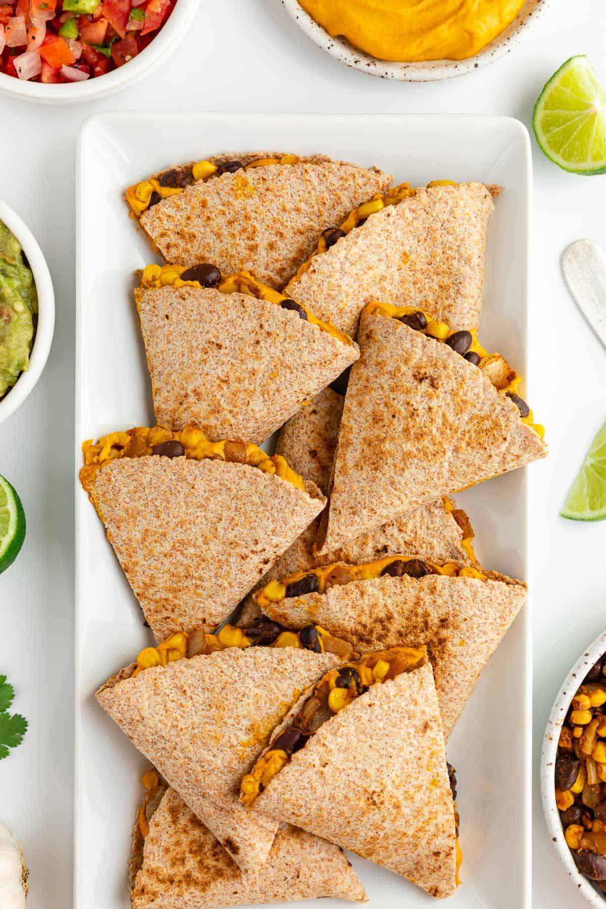 sweet potato black bean quesadillas on a white plate surrounded by bowls of guacamole, salsa, pico de gallo, and cilantro