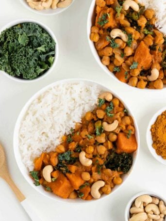 two bowls of sweet potato, kale, and chickpea curry beside a wooden spoon, bowl of kale, and bowl of cashews