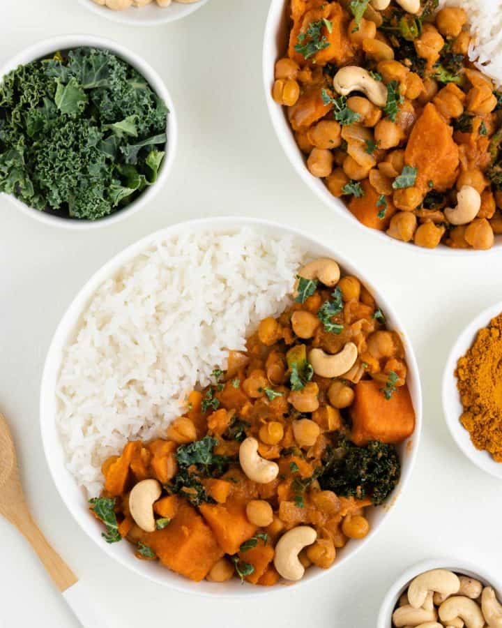 two bowls of sweet potato, kale, and chickpea curry beside a wooden spoon, bowl of kale, and bowl of cashews