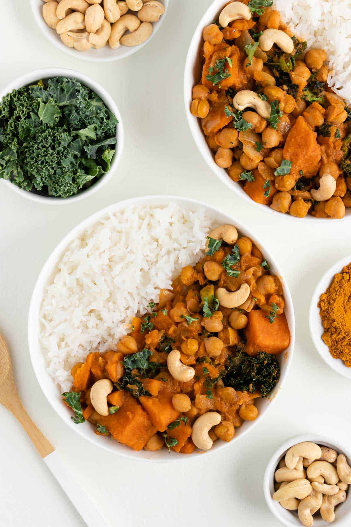 two bowls of sweet potato, kale, and chickpea curry beside a wooden spoon, bowl of kale, and bowl of cashews